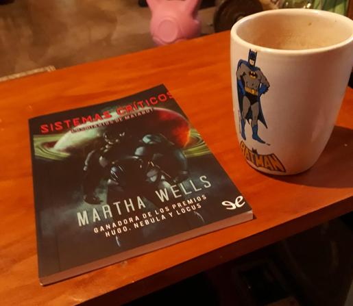 Día de lluvia, estoy leyendo "Sistemas críticos" de Martha Wells. Mi libro sobre la mesa junto a mí taza de café de batman .