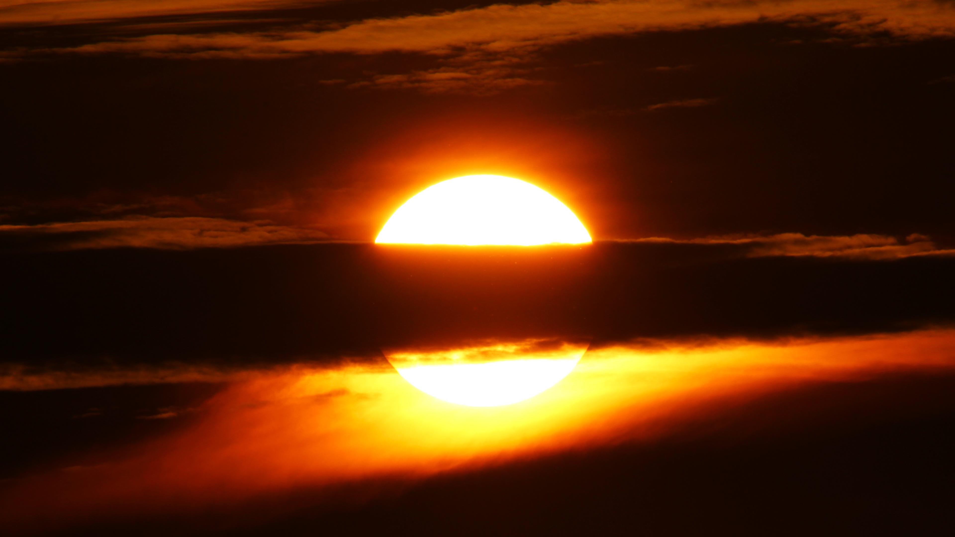 Sunset photo of a large white sun with a dark horizontal cloud across its middle, cutting it into a top and bottom part. The bright sunlight is picked up in several wispy clouds, especially below the dark horizontal cloud.