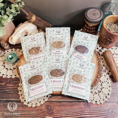Six small pouches of dried herbs are arranged on a wood surface. The pouches are about the size of a seed packet and have a clear window on the front. The label on each pouch features the name of herb it holds and its potential magickal uses, including angelica, wood betony, juniper berries, mistletoe, mullein, and horehound. This is an exclusive product from Inked Goddess Creations.