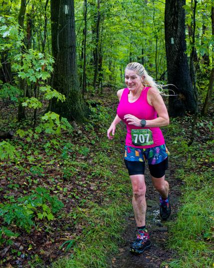 A woman with blond hair and a pink tank top has a green bib that say 707 in white is running on a path between two trees with rectangular white blazes