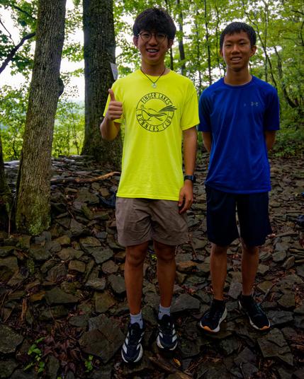 Two asian teenagers,  both wearing shorts and t-shirts and sneakers standing on a pile of flat shalely rocks with an open field behind a thin layer of trees.  One the left one is wearing a yellow FLRC volunteer shirt and is giving a thumbs up,  his cousin on the right is wearing a blue shirt.