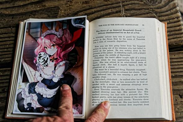 A book open on a wood plank floor with notable grain;  on the left find a bookmark picture of a beautiful woman with yellow eyes and red hair sitting on the edge of the outside floor of a Japanese house in a made outfit with fox ears and tail and a fan that says たまも(tamamo) and has a little doodle of a fox on it;  on the left find a story printed in letterpress with the header "The Story of An Imperial Household Guard Office Disillusioned by an Act of a Fox"
