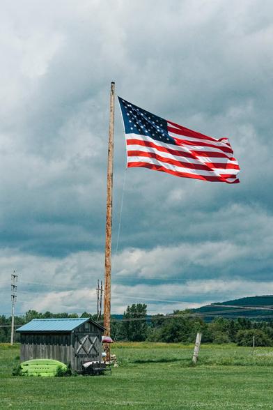 American flag with an impressive wood flagpole flying under a dramatic sky mostly filled with clouds with power lines behind  and a shed at the bottom over a close-cropped lawn