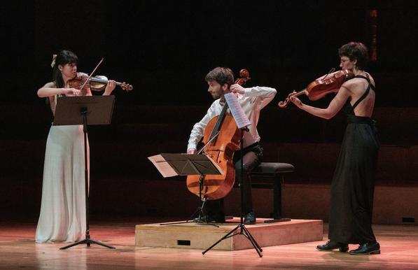 Lia, Marc et Coline en train de jouer de leurs instrument (respectivement violon, violoncelle et alto), sur la scène du De Singel à Anvers, lors de la finale du concours Supernova. Marc est tourné vers Lia, qui le regarde aussi. Les trois sont habillées en noir et blanc.
