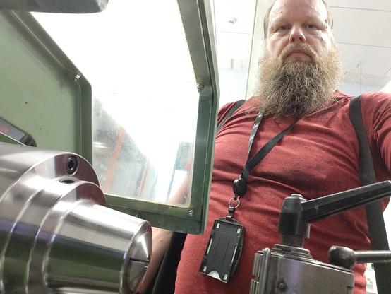 Parrakas punapaitainen mies vihreän sorvin ääressä.  Kuva otettu sorvin takaa.