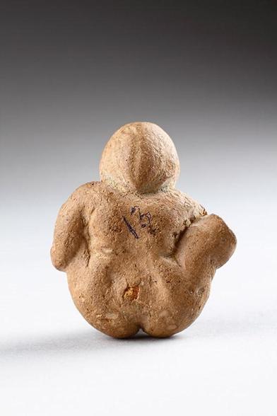 A votive terracotta of a woman sitting with her legs spread, seen from the reverse side. Her hair seems to be gathered but no bun is visible. The bum crack has been shaped with love for detail.