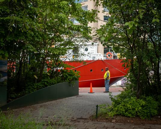 Color photo of a distant man in jeans, a long sleeve shirt, a hi-viz safety vest, and a trucker cap. He is framed right and left by some trees in a park, a brick path leads up between the trees to him. He is facing to the left, a single traffic cone stands before him. Beyond, a portion of the red hull and white superstructure of the Chesapeake lightship in Baltimore’s Inner Harbor forms the background.