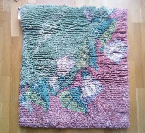 Laying flat on the floor, Finnish style handmade ryijy, but design is very modern. It has lots of colours, different shades of green on upper left corner, different shades of pink in lower right corner, wine of flower shapes and leafs going on diagonal accross it.  It looks vefy soft and fluffy, 
dimensions are ca 80cm x 1m