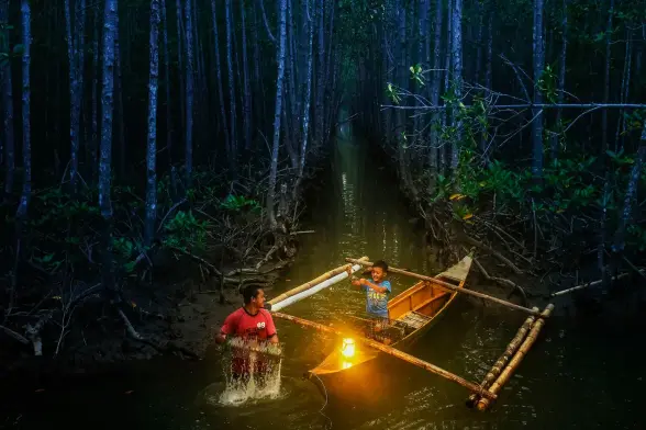 A child in a catamaran-like boat, while his father fishes with a net by the light on the bow.