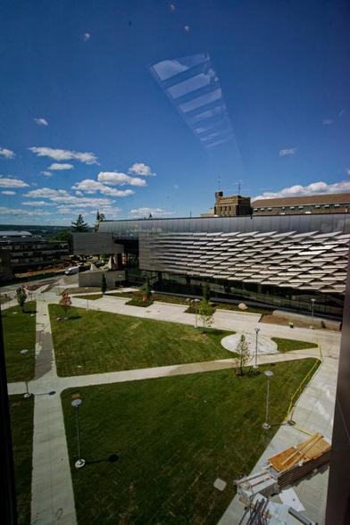 In the lower part of the picture find a grassy squard divided by two cement paths,  the paths are wet because they've been recently sprayed down, past some planters find a building with a large number of angled metal panels in front of and past that just a bit of a tall brown building with rows of windows,  across the street to the left edge is another building and above it all a blue sky with cumulus clouds in which a reflected line of windows is visible.