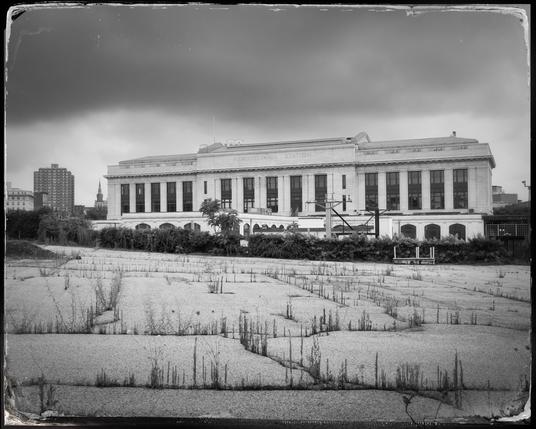 A black and white photo showing the back of Baltimore’s Penn Station, a large granite building, with a long closed off parking lot in the foreground. The parking lot’s pavement has many cracks through which scrubby grass is growing. Above the scene is a dark and foreboding sky.