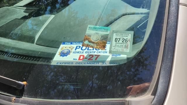 Front windshield of a car displaying decals from the "New York State Police investigators association" and a New York State safety inspection sticker. On the dashboard is a faded placard that reads "police identification d27"