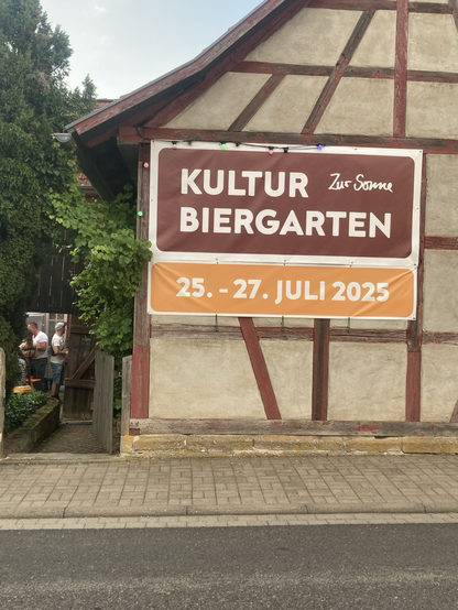 An einer Fachwerkscheune ist ein großes Banner angebracht. Darauf steht in weißen Buchstaben auf braunem Grund: Kulturbiergarten. Darunter weiß auf gelb: 25. bis 27. Juli 2025.