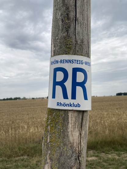 Auf einem Holzpfosten ist ein weißes Schild mit blauer Schrift angebracht. Darauf steht: Rhön-Rennsteig-Weg. RR. Rhönklub. Im Hintergrund sieht man ein Getreidefeld.