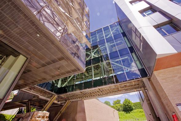Upward-facing view of a three-story bridge crossing from a new building on the right with vertical columns of windows divided with metal prisms and a glass and steel building on the left with perforated panels covering the windows