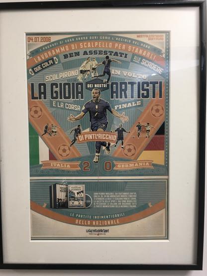 The image is a framed poster celebrating a soccer match between Italy and Germany, dated 04.07.2006. The poster features a central figure in a blue soccer uniform with the number 7, celebrating a goal. The text "LA GIOIA E LA CORSARA FINALE" is prominently displayed, along with "ITALIA 2 - 0 GERMANIA," indicating Italy's victory. The poster includes phrases like "I PROPRIONI DI CASA ERANO DURI COME L'ACCIAIO DEL NORD," "LAVORAMMO BEN ASSESTATI," and "ALLO SCADERE," reflecting the match's intens…