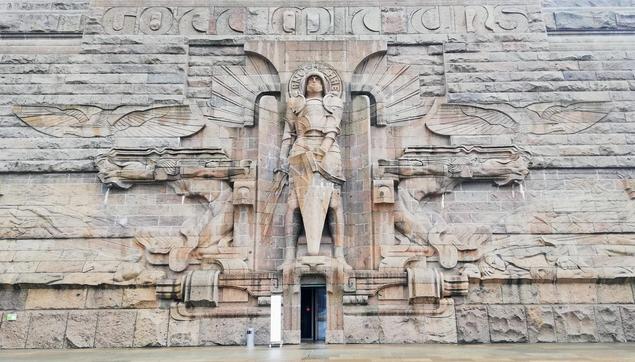 Über einer offenen Eingangstüre ist die riesige Wandstatue eines bewaffneten Engels, etwa 3 mal so hoch wie die Tür.