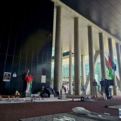 Wake voor Gaza bij het ministerie van Buitenlandse Zaken in Den Haag. Op de foto staat een man achter een microfoon met een Palestijnse vlag om de lessenaar, terwijl hij namen voorleest van door Israël vermoorde Palestijnen. Op de grond liggen matten, kussens, waterflessen en bloemen. Op de achtergrond zijn mensen aanwezig bij de wake en is de Palestijnse vlag zichtbaar. De wake vindt 24/7 plaats onder de overkapping van een modern gebouw met hoge zuilen.