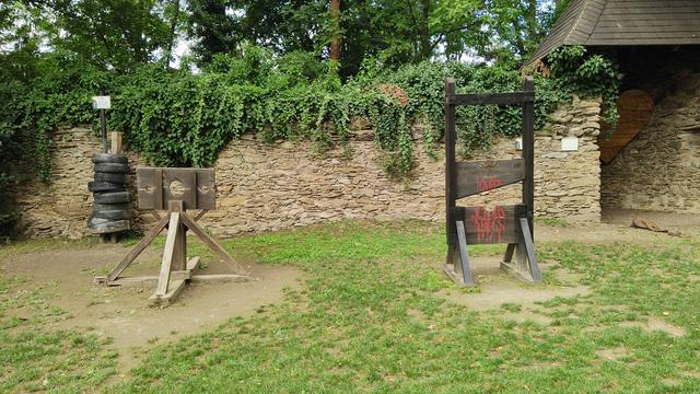 Auf einer grünen Wiese vor einer alten Mauer stehen links ein Pranger (Block) und rechts eine Guillotine, beide aus Holz. 
