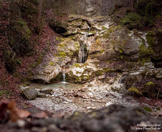 A small waterfall flows gently in a lush forest setting. The image showcases the serene beauty of nature, with the waterfall cascading over rocks and moss-covered surfaces. The surrounding trees provide a picturesque backdrop, creating a peaceful and tranquil atmosphere.  This landscape captures the essence of an outdoor oasis, perfect for those seeking a moment of relaxation and connection with the wilderness. The image exudes a sense of calmness and beauty, making it a perfect representation …