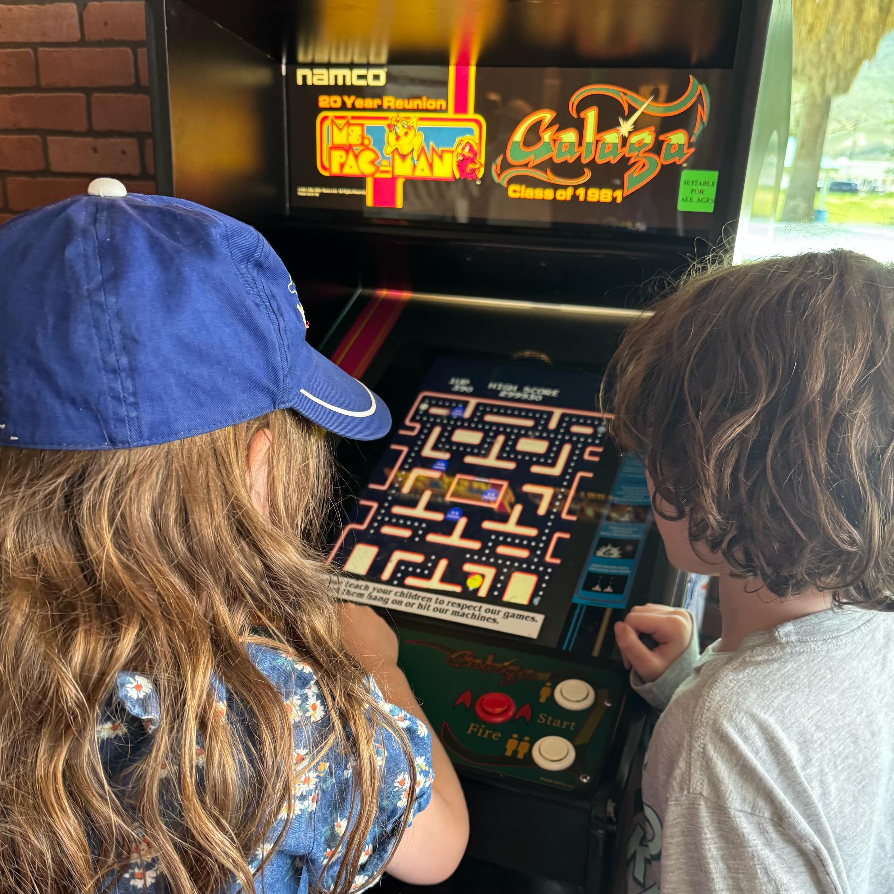 Two children are closely engaged in playing a classic arcade game on a retro-style machine. The cabinet features “Ms. Pac-Man” and “Galaga: Class of 1981” as part of the Namco 20 Year Reunion edition, with bright, nostalgic graphics on the screen and marquee. The child on the left has long hair, wears a blue cap and patterned shirt, and is actively using the joystick. The child on the right, with curly brown hair and a light-colored shirt, watches intently. The arcade screen shows the familiar maze layout of Ms. Pac-Man with colorful ghosts and dots, while the control panel includes red and white buttons labeled “Fire” and “Start.” The setting appears to be an indoor arcade or restaurant with natural light and outdoor greenery visible through a window in the background.