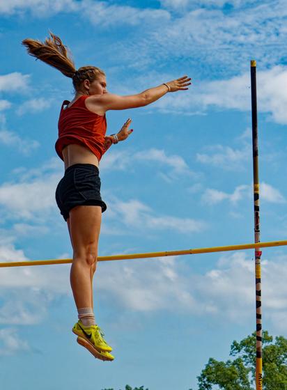 A woman hangs in the air perfectly vertically as she falls on the other side of the crossbar from the pole in the pole vault;  her twintails point up diagonally behind her head and her arms are reach out,  her organge tank top is floating up and you can see her back, she's wearing black sorts and yellow shoes and there is a complex cloudscape in the background