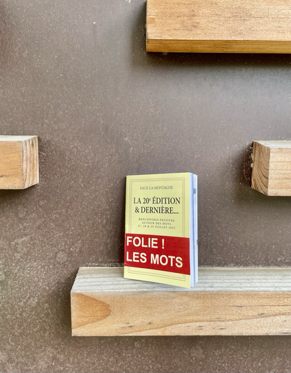 Photo du programme de la 20eme et dernière édition du festival Folie les Mots a Faux la Montagne. Maquette aux couleurs de la collection pages blanches de Gallimard. Le livret est posé sur un tasseau de bois, deux autres tasseaux pointent à droite et à gauche sur fond béton brun