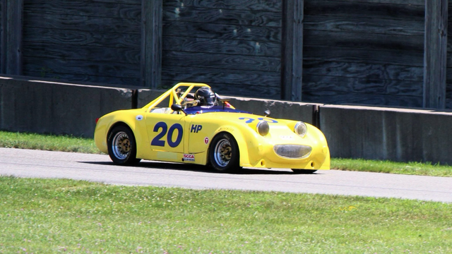 Photograph of a yellow “bug-eye” Austin-Healey Sprite seen from the side traveling to the right on a racetrack with grass in front and a retaining wall behind. The headlights and grill form its classic smile.