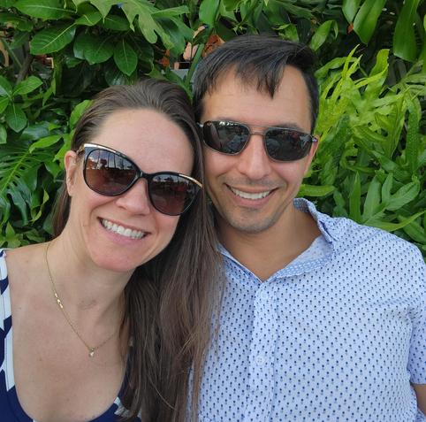 A man and woman, outside in front of greenery. Both are wearing sunglasses and smiling