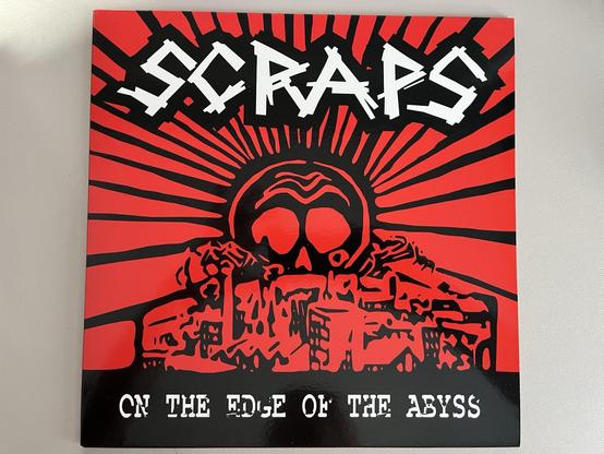 Das Bild zeigt ein Albumcover mit dem Titel "SCRAPS ON THE EDGE OF THE ABYSS". Das Design umfasst fette schwarze und weiße Buchstaben vor einem roten Hintergrund mit Strahlen, die von einem Schädel und einer Stadtlandschaft ausgehen, was ein auffälliges und dramatisches Bild ergibt.