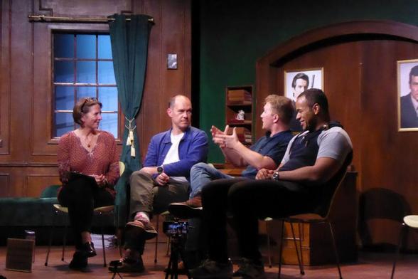 Four people seated on a stage, talking, with a wood-panelled stage set behind them.
