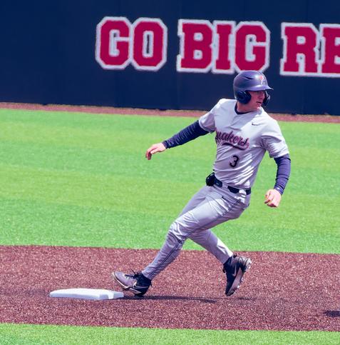 A baseball player reaches second base (I think) while looking back over his shoulder:  he's got a dark blue helmet and undershirt and a grey jersey that says Quakers 3 and behind him a sign reds GO BIG RED