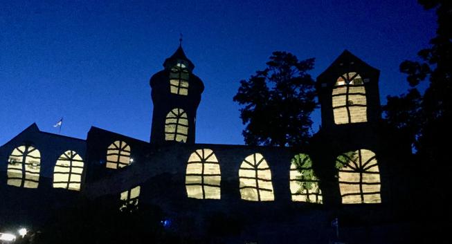 Vor einem tiefblauen Nachthimmel und mit einem Laubbaum im Hintergrund in der rechten Hälfte des Bildes hebt sich die Silhouette der Burg ab. Man sieht jedoch nicht die tatsächliche Baulichkeit, sondern eine vorgeblendete Projektion. Sie passt sich exakt den Umrissen des Gebäudes an und gibt die Illusion eines Scherenschnitts. Das Gemäuer ist schwarz, die angedeuteten Rundbögenfenster (sie stimmen nicht überein mit der Wirklichkeit) alle hell "beleuchtet" durch ganz leicht geld getöntes Licht. Bei genauem Hinsehen erkennt man hinter der Projektion Details wie Fahmen oder Äste, die sich gegen das projizierte Licht durchsetzen können. Unten links habe ich das Bild so beschnitten, dass die Straßenbeluchtung nur noch als kleiner Fleck zu erkennen ist.
Das Bild wirkt (zumindest auf mich :-)) märchenhaft.
