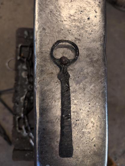A black, hand forged bottle opener made of steel. There are hammer marks running across the whole length of the handle leading to a round opening and a flattened piece to catch the lip of the bottle cap. 