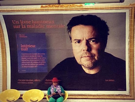 Dans le métro parisien, une grande affiche présentant le livre Intérieur nuit de Nicolas Demorand. Devant l'affiche, sur des sièges jaunes, est assise une femme vêtue de vert et portant un chapeau rouge. © #Off2Europe #VendrediLecture Gilles Denizot