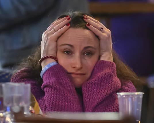 Middle-aged Woman with long puffy hair in a purple sweater holds her head,  there are a few plsatic cups in front of her