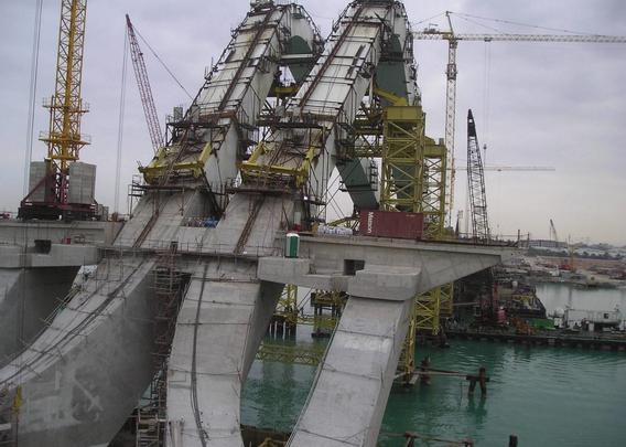 a photograph of a large construct of steel and concrete, it curves upward, two large concrete strips, water underneath, many cranes around it 