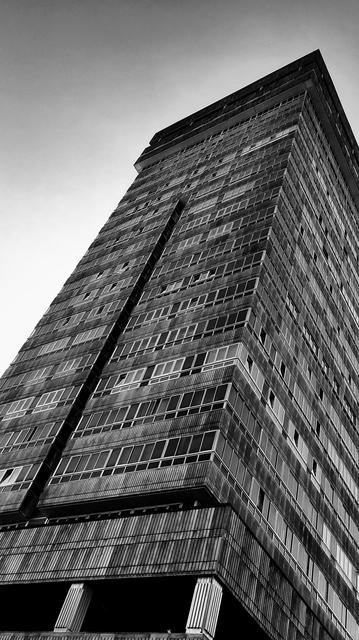 Schwarzweißfotografie: Ein vielstöckiges (ca. 15) Gebäude von schräg unten aufgenommen. 
Zwei Seiten sind zu sehen und zahlreiche Fenster erstrecken sich von unten bis oben.
