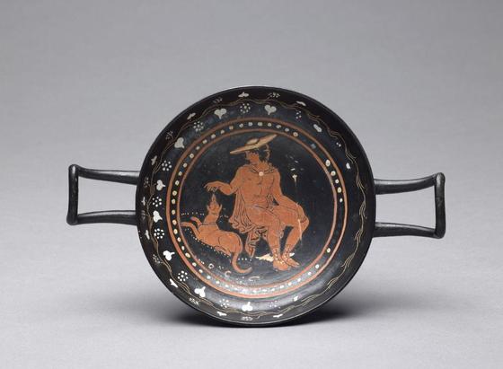 A delicately painted seated figure of Hermes adorns the centre of the kylix cup. Hermes feeds or playfully teases the hound reclining at his side. He wears his travelling hat and chlamys cloak. He's holding his kerykeion staff in his left and engaging with the hound with his right.