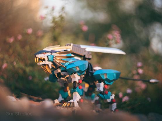 A Murasame Liger Zoid stands in a garden setting, its teal and gold armor contrasting with the soft pink flowers in the background. A single sword is mounted along its back, emphasizing its sleek, agile design. The robotic lion’s fierce expression and dynamic posture convey readiness for battle.