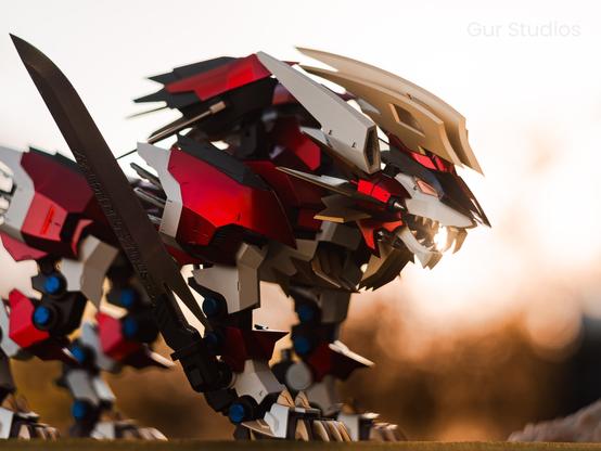 Close-up of a Hayate Liger Zoid, a mechanical lion with sleek red, gold, and silver armor plating. Its angular head is detailed with sharp fangs, vents, and blade-like fins, giving it a fierce, futuristic appearance. A sword is visible in the foreground, adding to the dynamic composition.