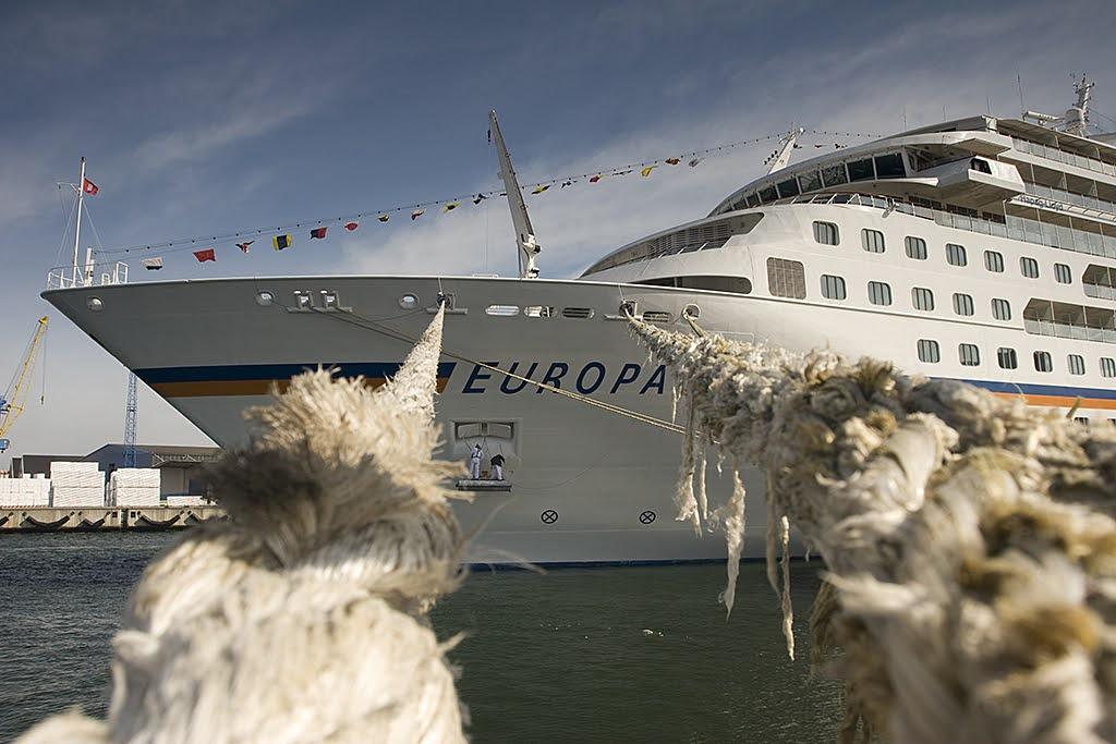 Bildbeschreibung von KI:
Das Foto zeigt ein großes Kreuzfahrtschiff mit dem Namen Europa, das am Kai eines Hafens festgemacht ist. Der Blickwinkel ist von schräg vorn aufgenommen, sodass der Bug des Schiffes dominiert. Zwei dicke, leicht ausgefranste Trossen aus hellem Tauwerk verlaufen aus dem Vordergrund schräg ins Bild hinein und verbinden das Schiff mit dem Kai. Diese Seile sind stark in den Fokus gerückt und wirken sehr massiv. Das Schiff selbst ist weiß mit mehreren Decks voller rechteckiger Fenster. Auf dem oberen Deck wehen kleine, bunte Signalflaggen an einer langen Leine über das Schiff gespannt. Man erkennt zwei Menschen in weißer Kleidung, vermutlich Crewmitglieder, die sich auf dem unteren Deck an der Reling aufhalten. Im Hintergrund sieht man einige Hafenanlagen mit Kranen, gestapelten weißen Containern und Gebäuden. Der Himmel ist leicht bewölkt, aber größtenteils blau. Die Szene wirkt ruhig, das Schiff liegt still im Wasser.
Bildbeschreibung von KI:
Das Foto zeigt ein großes Kreuzfahrtschiff mit dem Namen Europa, das am Kai eines Hafens festgemacht ist. Der Blickwinkel ist von schräg vorn aufgenommen, sodass der Bug des Schiffes dominiert. Zwei dicke, leicht ausgefranste Trossen aus hellem Tauwerk verlaufen aus dem Vordergrund schräg ins Bild hinein und verbinden das Schiff mit dem Kai. Diese Seile sind stark in den Fokus gerückt und wirken sehr massiv. Das Schiff selbst ist weiß mit mehreren Decks voller rechteckiger Fenster. Auf dem oberen Deck wehen kleine, bunte Signalflaggen an einer langen Leine über das Schiff gespannt. Man erkennt zwei Menschen in weißer Kleidung, vermutlich Crewmitglieder, die sich auf dem unteren Deck an der Reling aufhalten. Im Hintergrund sieht man einige Hafenanlagen mit Kranen, gestapelten weißen Containern und Gebäuden. Der Himmel ist leicht bewölkt, aber größtenteils blau. Die Szene wirkt ruhig, das Schiff liegt still im Wasser.