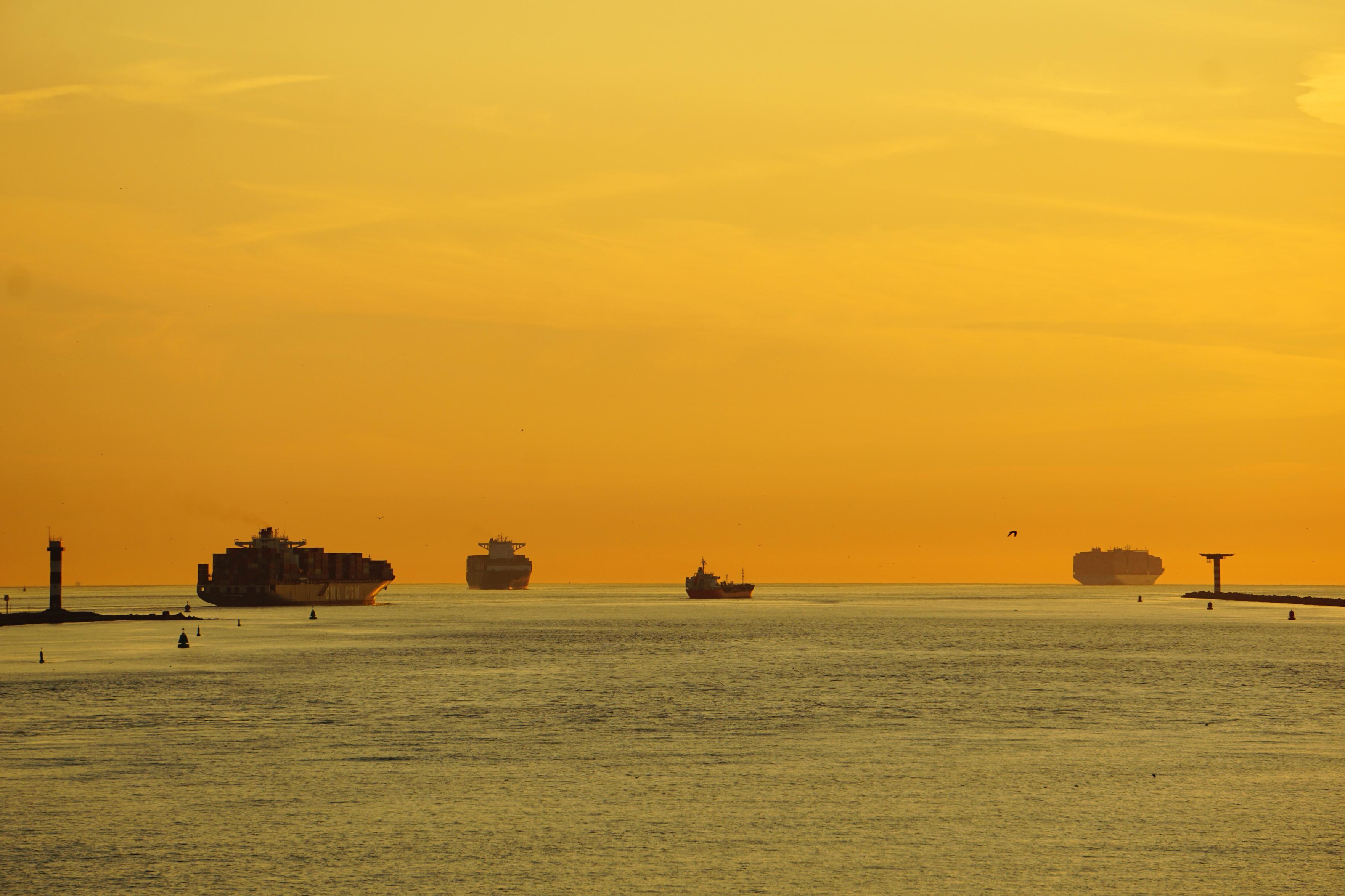 Blick auf das offene Meer vom Rotterdamer Hafen. Himmel und Meer sind durch die untergehende Sonne gelb gefärbt. Rechts und links laufen die Hafenmolen aus. Am Horizont sind drei Containerschiffe und ein kleinerer Frachter zu sehen. Blick auf das offene Meer vom Rotterdamer Hafen. Himmel und Meer sind durch die untergehende Sonne gelb gefärbt. Rechts und links laufen die Hafenmolen aus. Am Horizont sind drei Containerschiffe und ein kleinerer Frachter zu sehen.