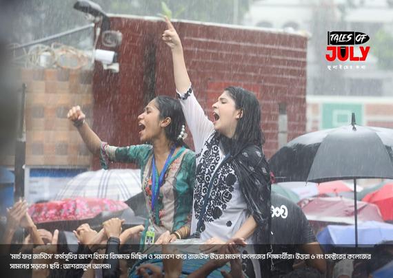 Two young women, soaked in heavy rain, passionately lead a protest while standing above a crowd holding umbrellas. One raises her fist and the other points upward, both shouting slogans. The atmosphere is intense and defiant. The event is part of the “March for Justice” movement involving students from Uttara Model College and Uttara 6 No. Sector College in Bangladesh. A red brick wall and surveillance camera are visible in the background, with the “Tales of July” logo in the corner.