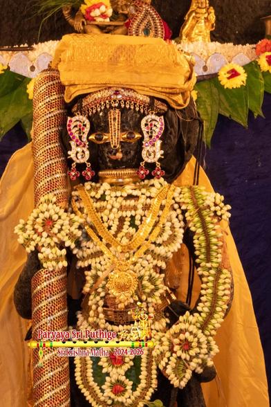 Closeup of idol of Hindu deity Krishna in yellow cape and turban standing visible only above the knees. A gilded (with orange striations) staff in right hand and left hand on front left waist. His waist and front down to knees as well as the entire length of the left arm, hand, and palm are lined with a long string of white jasmine buds. His upper body is also covered by a contouring string of jasmine flowers, placed to take the outline of a vest. A string of green beetle leaves in the background.