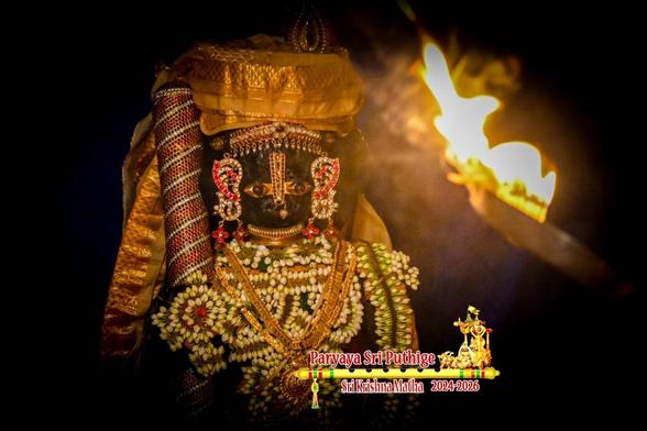 Closeup of idol of Hindu deity Krishna in yellow cape and turban with light (aarati) shown close in front of his face. Top of a gilded (with orange striations) staff visible in right hand. Only the left upper arm is visible. His left arm and his chest are lined with a long string of white jasmine buds.