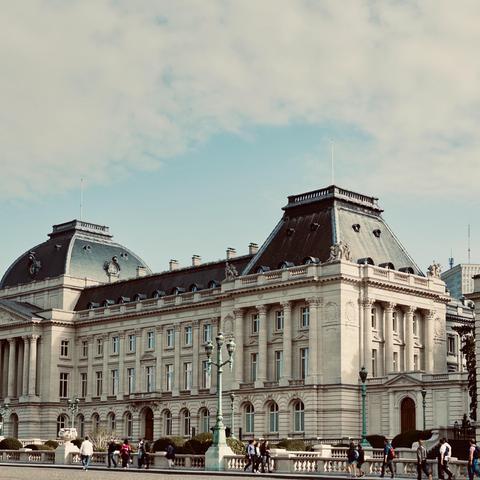 The image shows a large, elegant building with a classic European design. The building has light beige stone walls and a dark gray roof with steep slopes. There are many tall windows arranged in rows, with decorative frames and small balconies. The roof has round windows and sculptures near the edges. Two main sections of the building have high, square-shaped towers with flat tops and flagpoles. In front of the building, there are ornate green street lamps and stone statues. A group of people is walking on a wide pavement, adding life to the scene. The sky is partly cloudy with patches of blue.