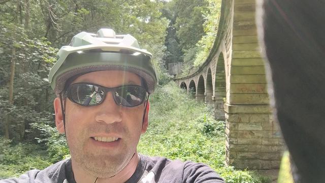 A guy in sports shades and a green cycle helmet smiling at the camera. Behind is an old curving aqueduct made of stone. 