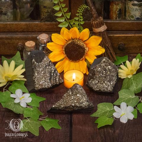 Three specimens of the "Pyrite with Shungite Raw Gemstone" product are arranged on a wooden surface. The pyrite sparkles in shades of gold and brown, contrasting with the matte black of shungite. Presented by Inked Goddess Creations.
