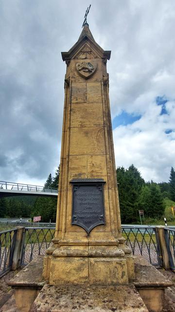 Ein etwa 7 Meter hoher Obelisk aus gelbem Stein. Eine dunkle Metalltafel mit Inschrift ist darauf angebracht. Im Hintergrund eine geschwungene Fußgängerbrücke.